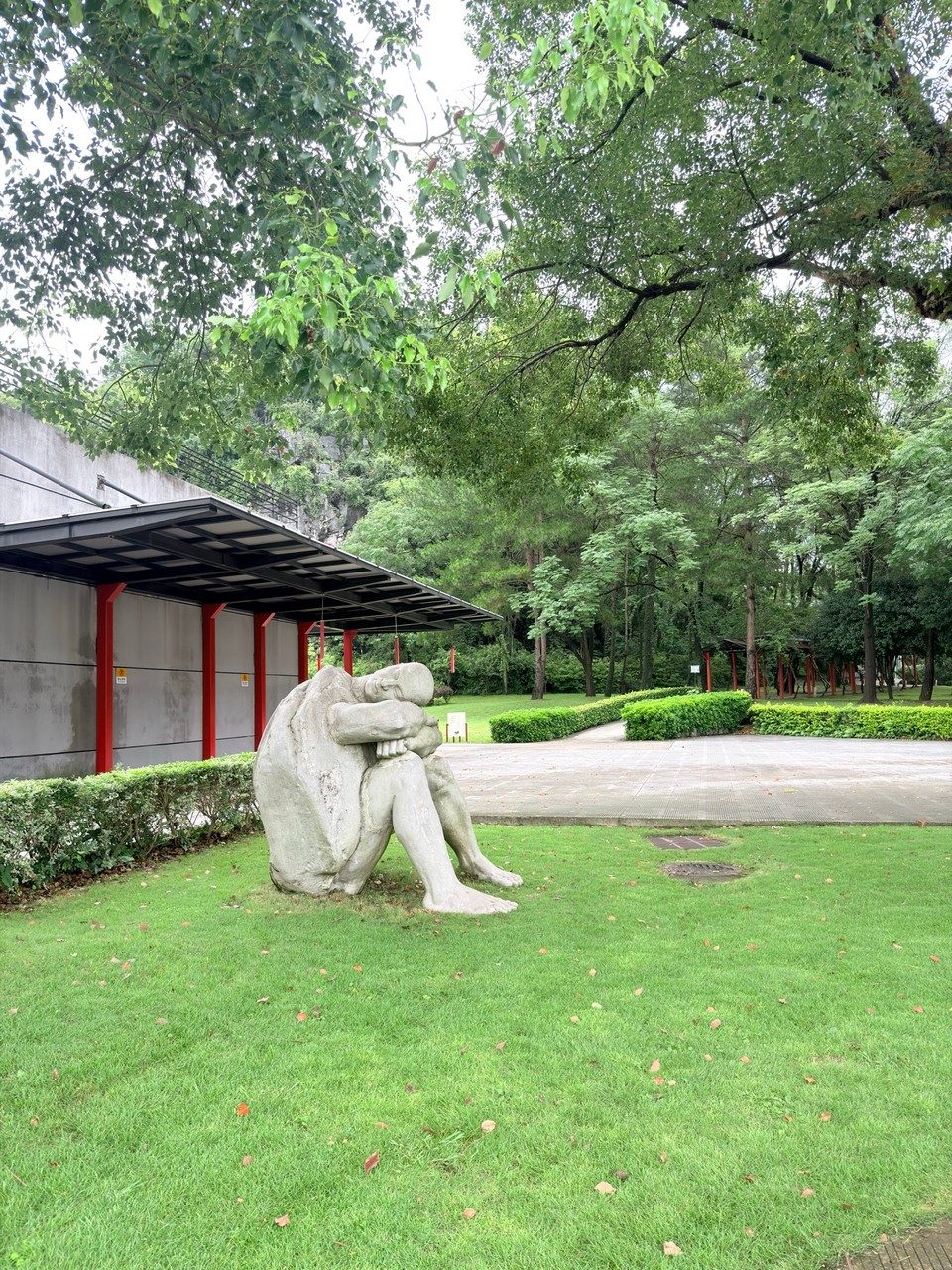 A white stone statue of a person reading, sitting on grass beside a tree-lined path