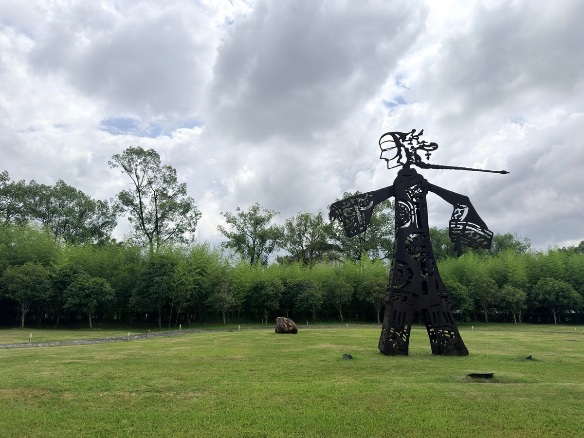 A tall steel sculpture of a figure standing on green grass under a cloudy sky